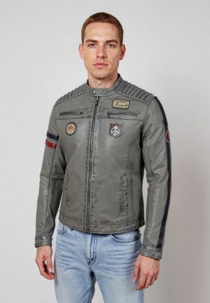 Junger Mann trägt eine graue Zip-up-Jacke mit Aufnähern und blaue Jeans, steht vor einem einfachen hellen Hintergrund.
