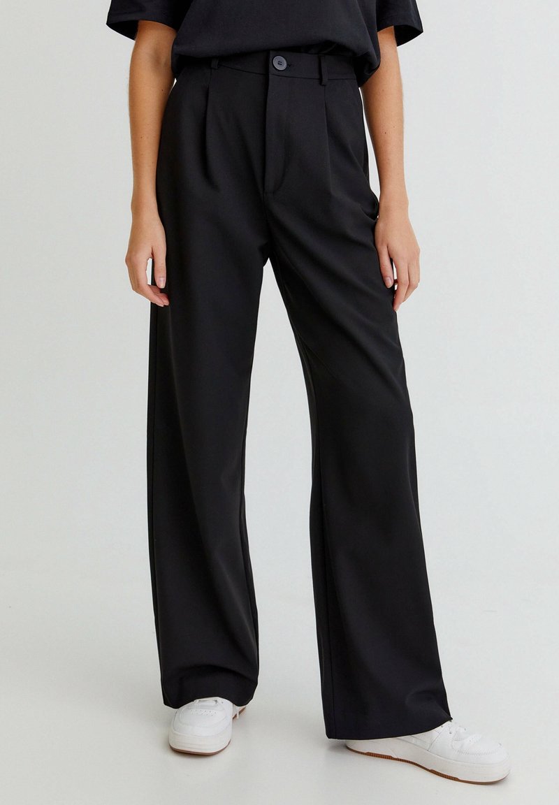 Personne portant un pantalon noir ample avec un bouton à l'avant, associé à des baskets blanches, se tenant devant un fond uni.