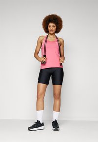 Débardeur de sport rose avec des bretelles fines, assorti à un short de compression noir. Corde à sauter noire dans les mains, sneakers noires et chaussettes blanches.