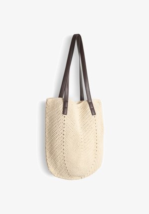 Sac cabas tissé de couleur crème avec de longues anses en cuir brun foncé suspendu devant un fond blanc.