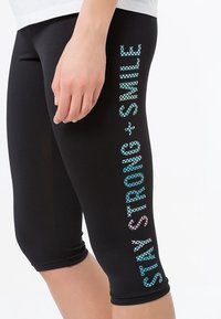 Leggings neri con un design aderente, lunghezza cropped e una stampa grafica colorata con scritto "STAY STRONG + SMILE" lungo il lato.