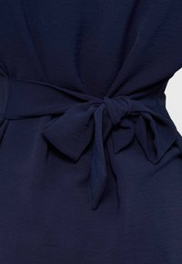 Υφασμάτινο σε ναυτικό μπλε δεμένο σε φιόγκο στη μέση ενός ενδύματος, παρουσιάζοντας υφή και πτυχώσεις.