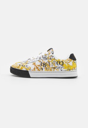 Biały niski sneaker z żółtymi barokowymi wzorami kwiatowymi, czarnym tekstem, białymi sznurówkami oraz czarnym brandem na podeszwie na jednolitym tle.