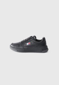 Black leather sneaker with a textured surface, flat sole, and lace-up design. Features a logo patch on the side in blue, red, and white colors.