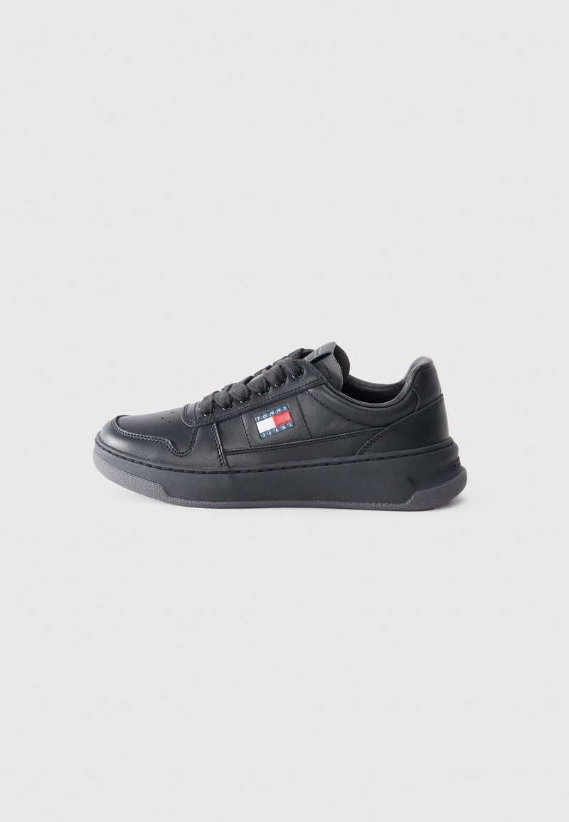 Black leather sneaker with a textured surface, flat sole, and lace-up design. Features a logo patch on the side in blue, red, and white colors.