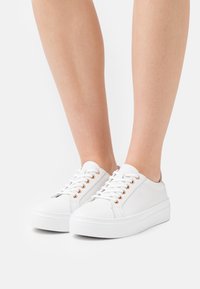 Des baskets blanches en cuir présentent une chaussure à bout rond, une semelle plate et six œillets avec des éléments en or rose. Texture lisse avec des détails de design minimaux.