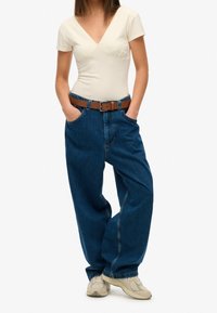 Cremefarvet tætsiddende top med korte ærmer, parret med løse blå denimbukser og et brunt læderbælte. Hvide sneakers fuldender outfittet.