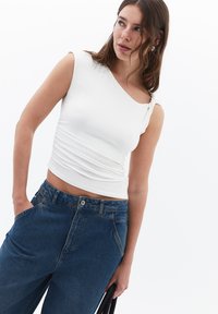 Bílé bezramenní tričko s jedním ramínkem a nařasením, doplněné o high-waisted modré džíny.