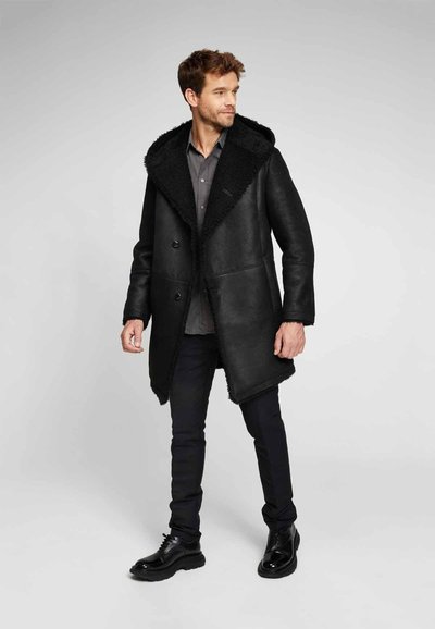 Cappotto in finta pelle nera con un ampio colletto in pelliccia testurizzata, chiusura a doppio bottone e una vestibilità leggermente oversize, abbinato a pantaloni neri e scarpe.