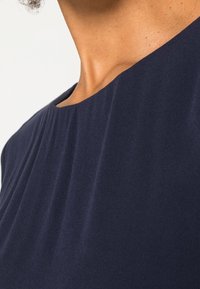 Primer plano del cuello y la parte superior del pecho de una persona que lleva una prenda de cuello redondo de color azul marino oscuro, contra un fondo claro y liso.