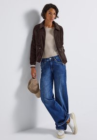Brun mockajacka med vita randiga manschetter, beige topp, vida blå jeans och grå sneakers. Håller en beige keps, mot en ljus bakgrund.