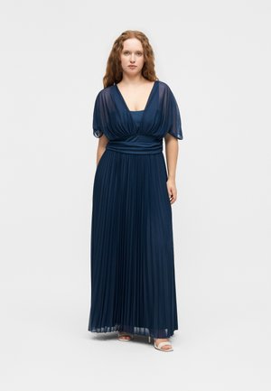 Femme aux longs cheveux bouclés portant une robe plissée bleu marine fluide avec des manches courtes transparentes et des sandales blanches à talons, debout devant un fond uni.