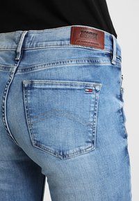 Vaqueros de mezclilla azul claro con un parche de cuero marrón que dice "TOMMY JEANS." Presenta un bolsillo trasero con detalle de costura y un acento de color.