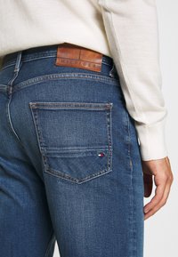 Close-up of person wearing blue Tommy Hilfiger jeans with brown leather brand patch and small logo on back pocket, paired with cream sweater.