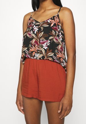 Kobieta ubrana w czarny top bez rękawów z kolorowym, kwiatowym wzorem oraz rdzawobrązowe szorty z wysokim stanem na gładkim tle.