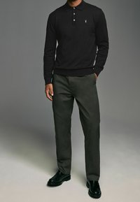 Zwarte polo met lange mouwen en drie knopen, voorzien van een klein embleem, gecombineerd met donkergroene broek en zwarte schoenen met gestructureerde zolen.