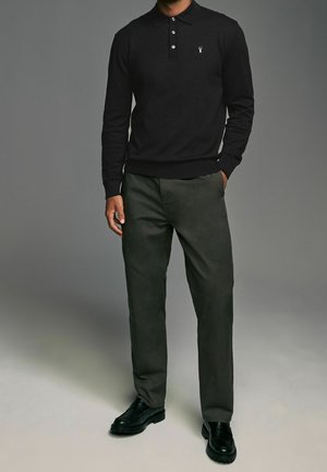 Schwarzes Langarm-Poloshirt mit drei Knöpfen und kleinem Emblem, kombiniert mit dunkelolivgrünen Hosen und schwarzen Schuhen mit strukturierten Sohlen.