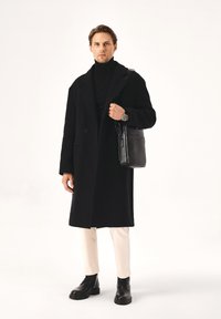 Cappotto di lana nero con ampi rever, abbinato a un dolcevita nero, pantaloni color crema e stivali neri con lacci, mentre tiene una borsa nera strutturata.