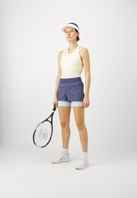 Κίτρινο αμάνικο μπλουζάκι, ναυτικό μπλε και ανοιχτό μπλε σορτς, κρατώντας μια μαύρη ρακέτα τένις. Λευκό καπέλο και παπούτσια με ροζ λεπτομέρειες. Απλός, αθλητικός σχεδιασμός.