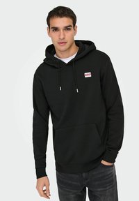Zwarte hoodie van zacht materiaal, met een voorzak en een klein rood label linksboven. Klassiek ontwerp met geribbelde boorden.