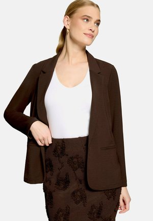 Femme portant un blazer marron foncé sur un top blanc à encolure dégagée et une jupe fleurie marron foncé texturée, cheveux tirés en arrière et boucles d'oreilles créoles dorées.
