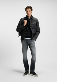 Svart puffjacka med knappstängning, två bröstfickor, slim-fit grå jeans och svarta sneakers med vita sulor, bärs över en mörk tröja.