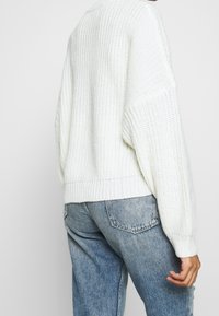 Personne portant un pull en grosse maille blanc et un jean bleu clair délavé, vue de dos avec une main visible.