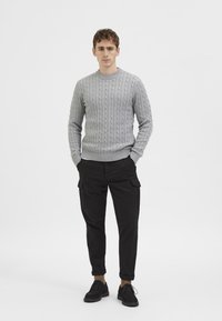 Szürke, kötött mintás pulóver, fekete oldalzsebes cargo nadrág, fekete cipő, sima fehér háttér előtt álló.