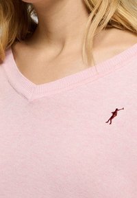 Light pink sweater featuring a v-neck design, made from soft fabric, with a small embroidered figure on the left chest.