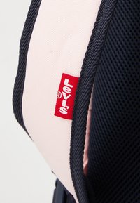 A Levi's hátizsák részlete, rózsaszín anyaggal és sötétkék háló díszítéssel, egy piros Levi's címkével jelölve, kiemelve a textúrát és a színkontrasztot.