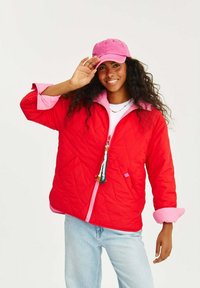 Červená prešívaná bunda s ružovým golierom a manžetami, so zapínaním na zips vpredu a dekoračným strapcom. Nosená s ružovou čiapkou a pohodlnými džínsami.