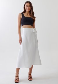 Jupe enveloppante blanche avec un détail de lien à la taille, associée à un crop top noir ajusté. La jupe est fabriquée dans un tissu lisse et fluide.