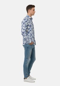 Joven con camisa azul de manga larga con estampado floral, vaqueros ajustados desgastados y zapatillas blancas, de pie de perfil con las manos en los bolsillos sobre fondo blanco.