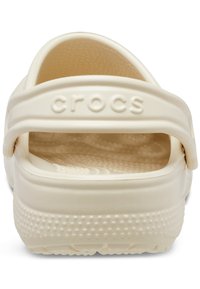 Beli Crocs obutev ima zaprt petni del, gladek plastičen material, drenažne luknje in napisano logotip na zadnjem traku. Ima zaobljeno obliko z teksturiranim notranjim delom.