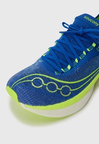 Blue athletic shoe with a mesh upper and neon green accents. Features laces, circular detailing, and a white sole with a slightly elevated heel.