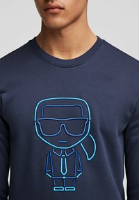 Man som bär en marinblå sweatshirt med blå linjekonstdesign av en person med solglasögon, slips och hästsvans på framsidan.