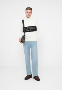 Balts rāvējslēdzēja džemperis ar melnu "ILFIGER Racing Club" logo, gaiši zili džinsi un brūnas kurpes. Melna plecu soma. Tīra, minimāla fona.