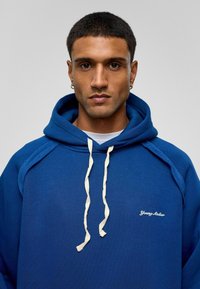 Blauwe hoodie gemaakt van zacht materiaal met een trekkoordsluiting. Heeft een wit geborduurd logo op de borst en een kenmerkend hoodie-ontwerp.