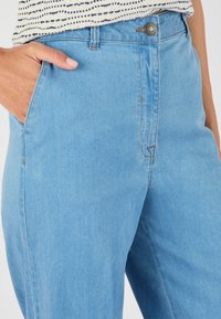 Pantalones de mezclilla azul claro con una cintura ajustada, bolsillos frontales y un cierre clásico de cremallera y botón. Presenta detalles de costura sutiles.