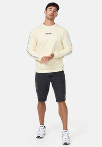 Sudadera de color amarillo claro con mangas largas, con texto negro en las mangas, combinada con pantalones cortos oscuros y zapatillas blancas.