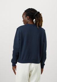 Námořnická modrá mikina s dlouhými rukávy, kulatým výstřihem a žebrovanými manžetami. Hladká textura, volný střih a minimální designové detaily.