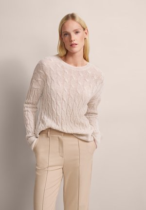 Rundhals-Pullover aus einem Off-White-Wollmix mit einem strukturierten Zopfmuster; kombiniert mit hellbeigen, taillierten Stoffhosen.