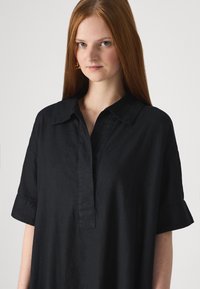 Camisa de linho preta com gola, mangas curtas e fecho frontal. O tecido tem uma aparência ligeiramente texturizada.