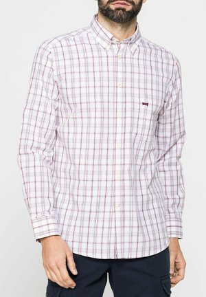 Uomo con camicia bianca a bottoni con motivo a quadri rossi e pantaloni scuri, in piedi davanti a uno sfondo semplice.