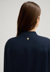 Marineblaue Bluse mit glatter Textur, ausgestattet mit einem Kragen und einem runden silbernen Knopfdetail auf der Rückseite in der Nähe des Halsauschnitts.
