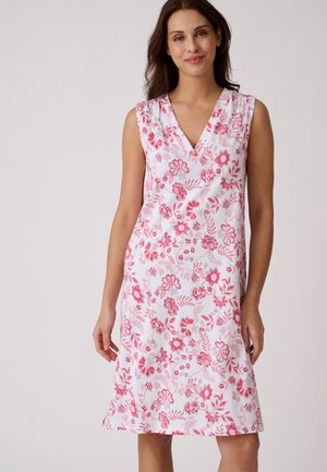 Femme portant une robe blanche sans manches avec des motifs floraux rouges et roses, debout devant un fond clair uni.