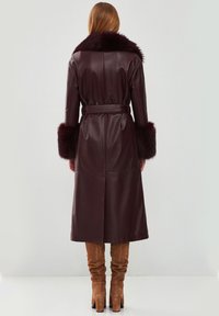 Cappotto lungo in pelle bordeaux con colletto e polsini in pelliccia, vita con cintura e taglio dritto. Presenta uno spacco sul retro per facilitare i movimenti.