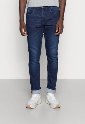 Mężczyzna ubrany w ciemnoniebieskie obcisłe dżinsy z mankietami i białe trampki, stojący na jednolitym jasnym tle.