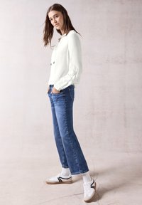Top bianco a maniche lunghe con tessuto testurizzato, jeans blu e sneakers bianche con dettagli neri. Modello in posa di lato su uno sfondo chiaro.