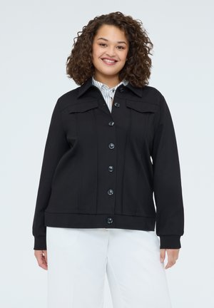 Uśmiechnięta kobieta o kręconych włosach, ubrana w czarną kurtkę zapinaną na guziki na paskowaną koszulę oraz białe spodnie, stojąca na tle jednolitego tła.